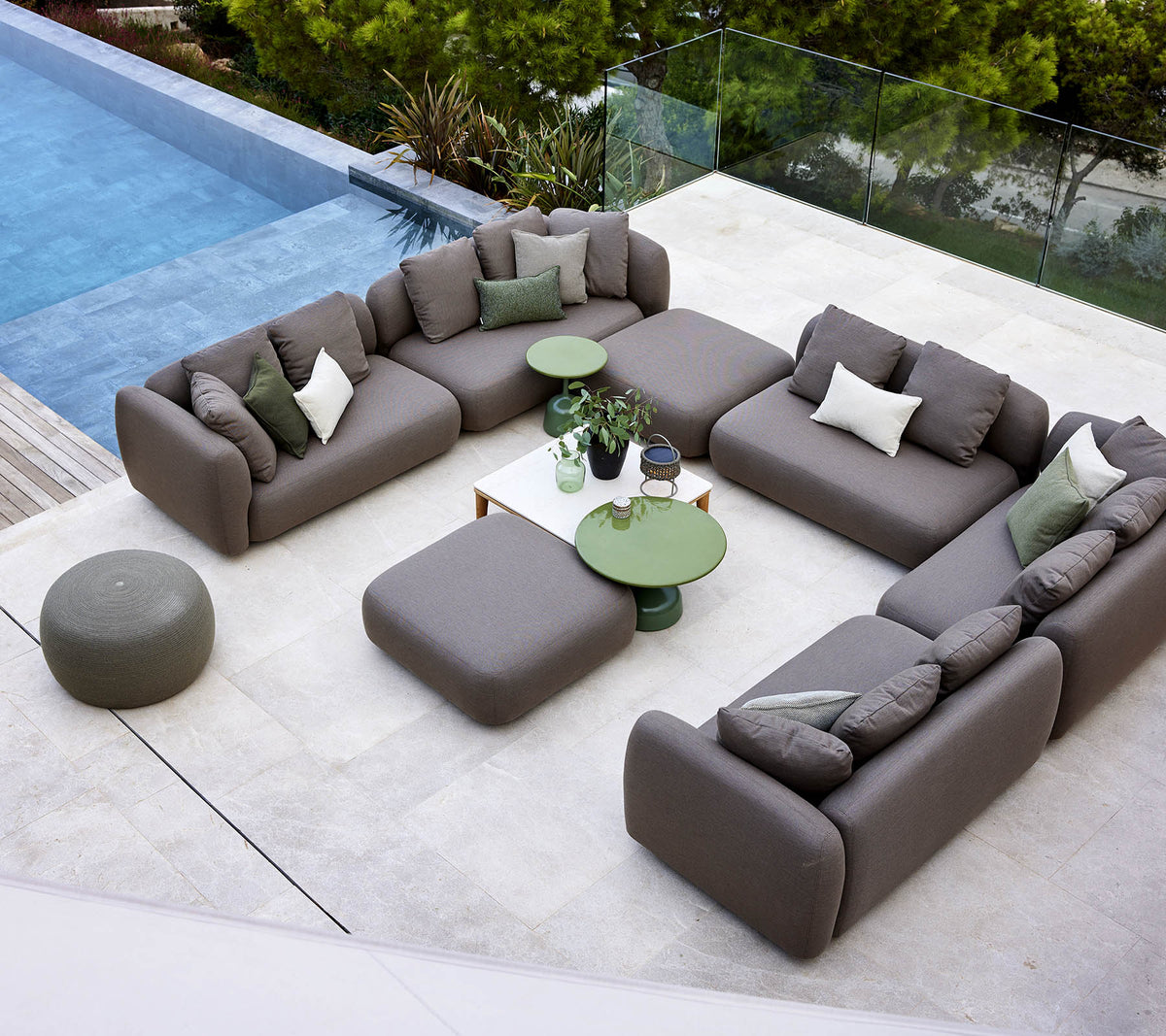 Eine moderne Lounge-Ausstattung mit einer Eckcouch, einem runden Ottoman und grünen Beistelltischen, umgeben von Grünpflanzen und einem ruhigen Pool.
