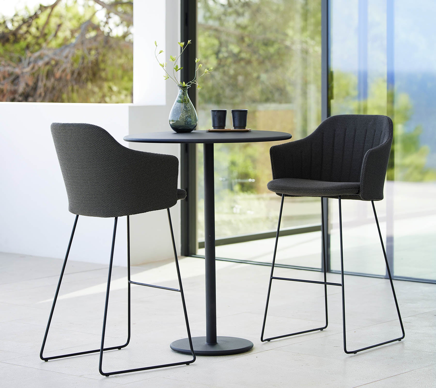 Zwei stilvolle Wahl-Barhocker, kombiniert mit einem schlanken runden Bartisch, akzentuiert durch dekorative Gegenstände und einen natürlichen Hintergrund.