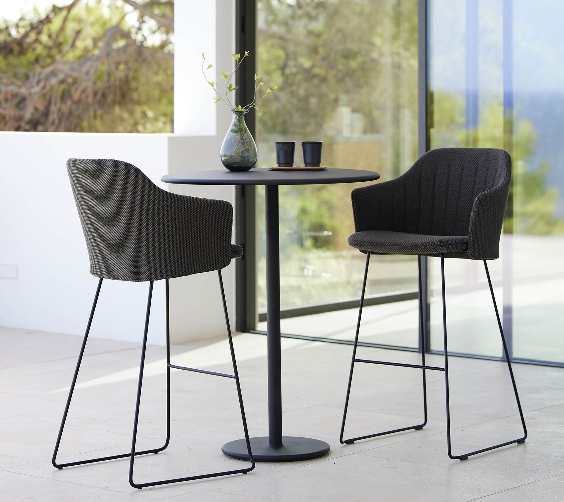 Zwei stilvolle Stühle kombiniert mit einem eleganten Bartisch, ergänzt durch eine Vase und Tassen, schaffen eine moderne und einladende Sitzgelegenheit.