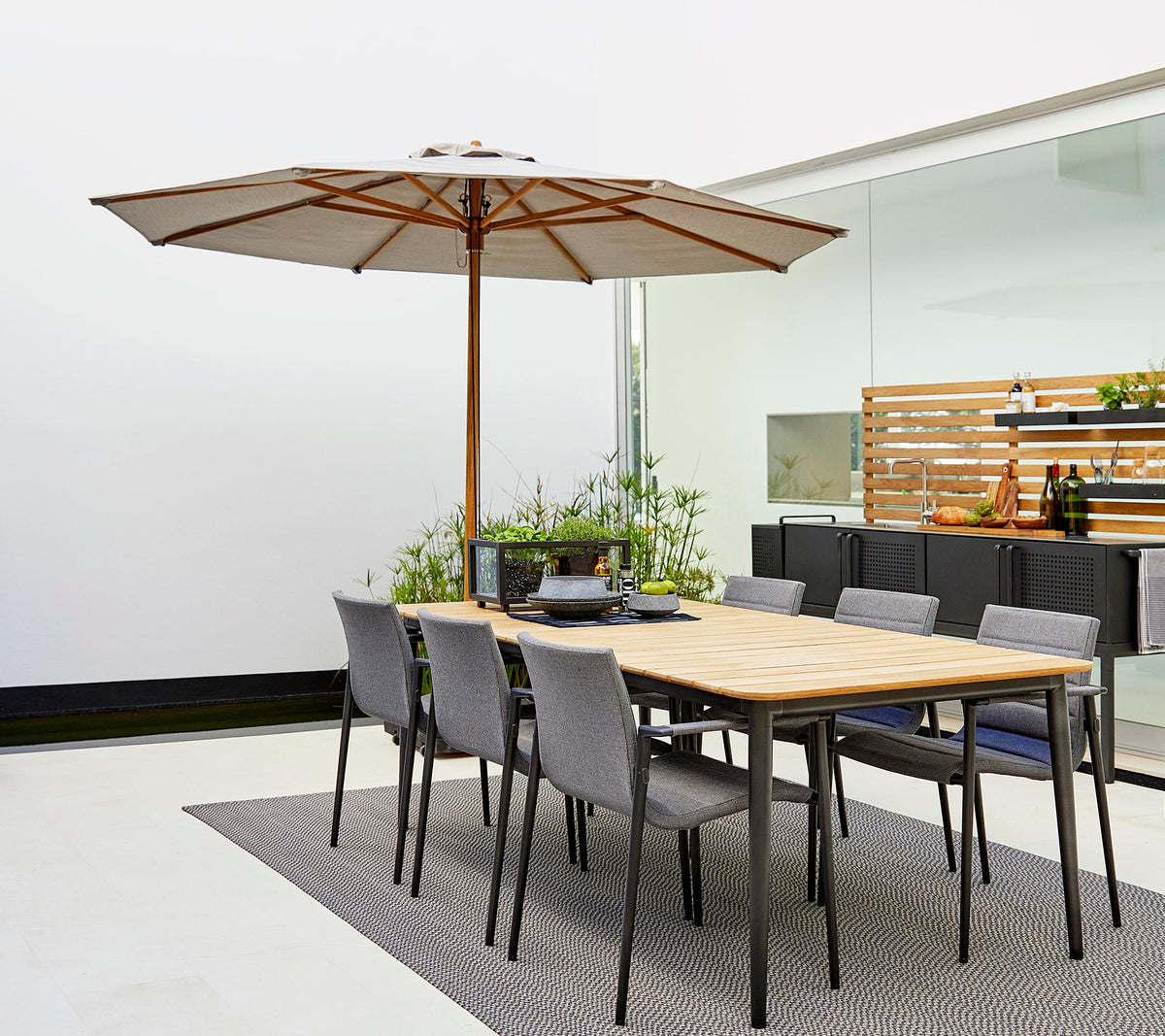 Ein klassisches Esszimmer-Set mit einem Holztisch mit einem Sonnenschirm und grauen Stühlen, umgeben von dekorativen Pflanzen auf einem stilvollen Teppich.