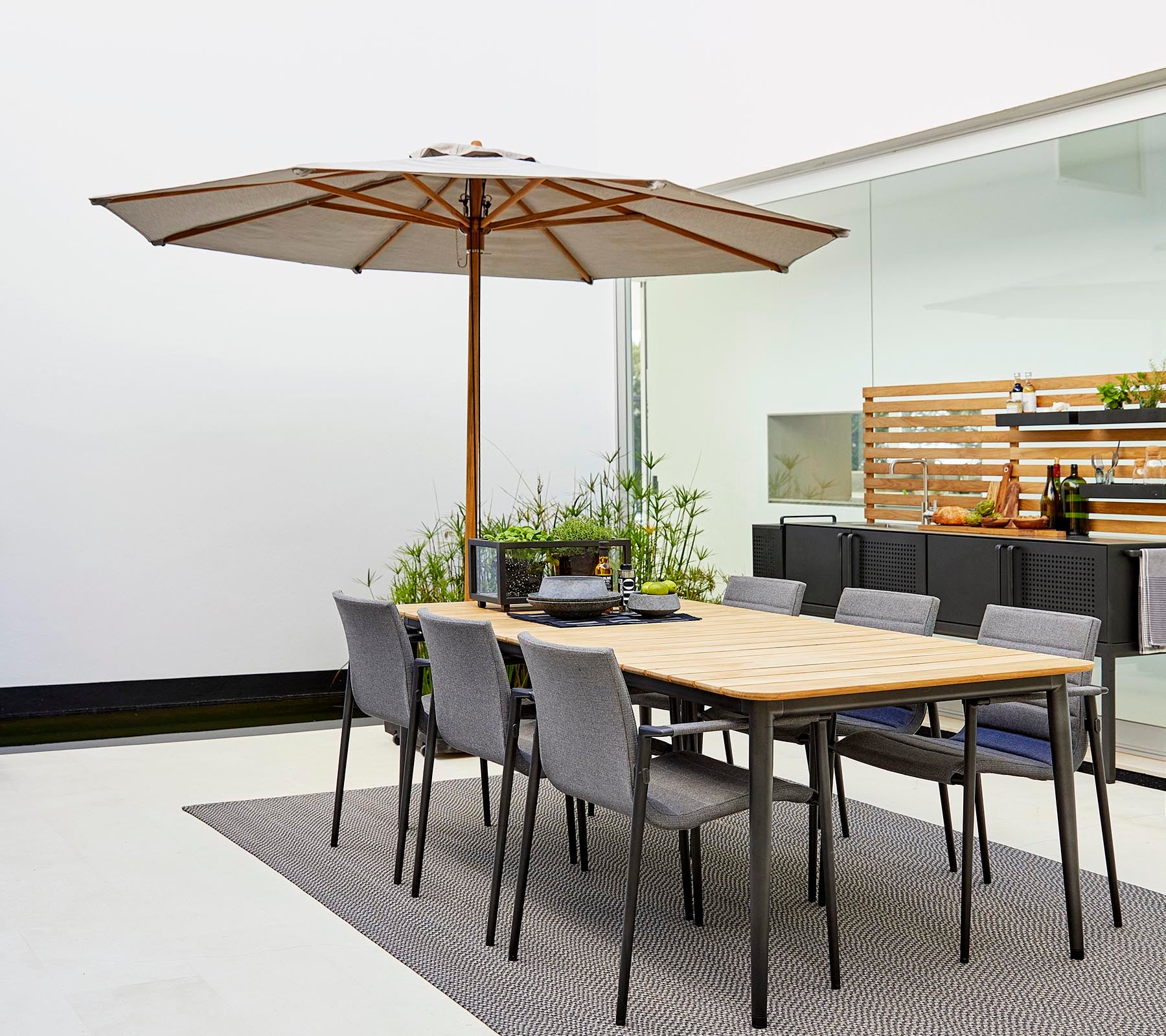 Ein klassisches Esszimmer-Set mit einem Holztisch mit einem Sonnenschirm und grauen Stühlen, umgeben von dekorativen Pflanzen auf einem stilvollen Teppich.