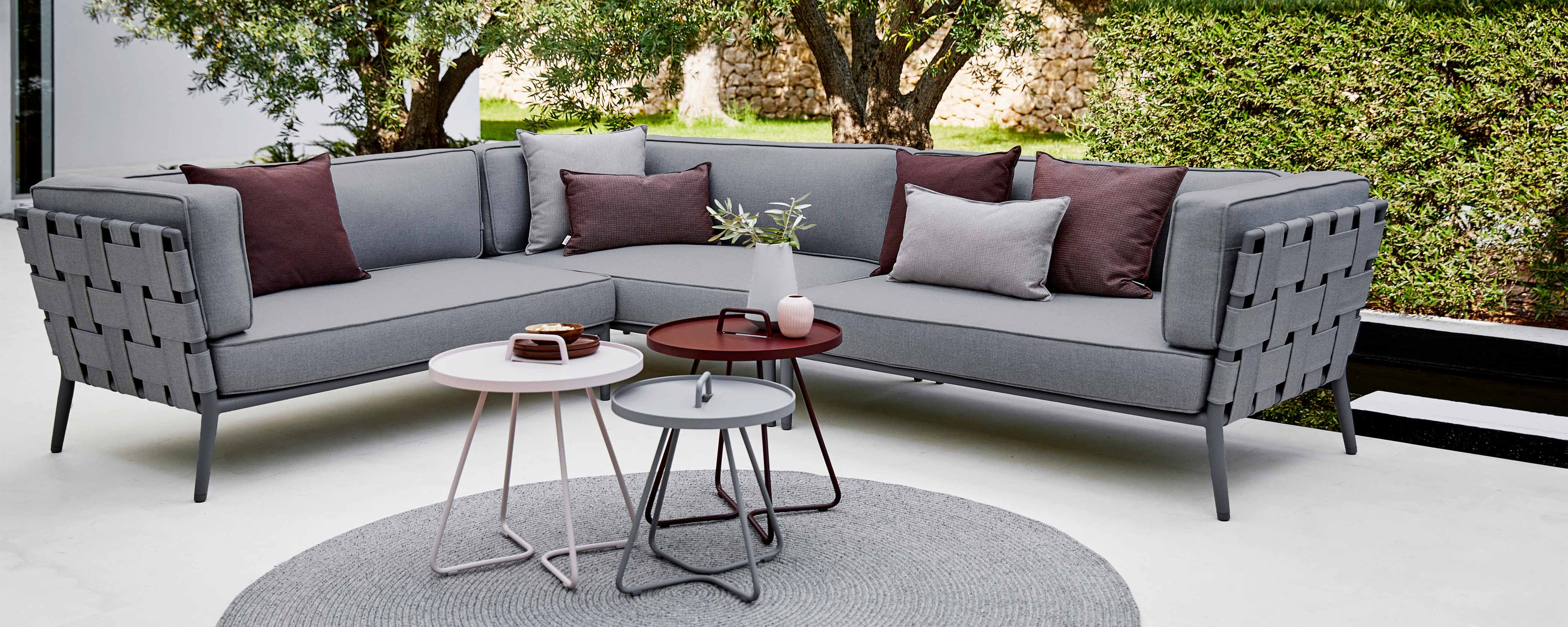 Moderne konische Ecksofa aus grauem Stoff mit dekorativen Kissen, begleitet von stilvollen Beistelltischen und einem runden Teppich.