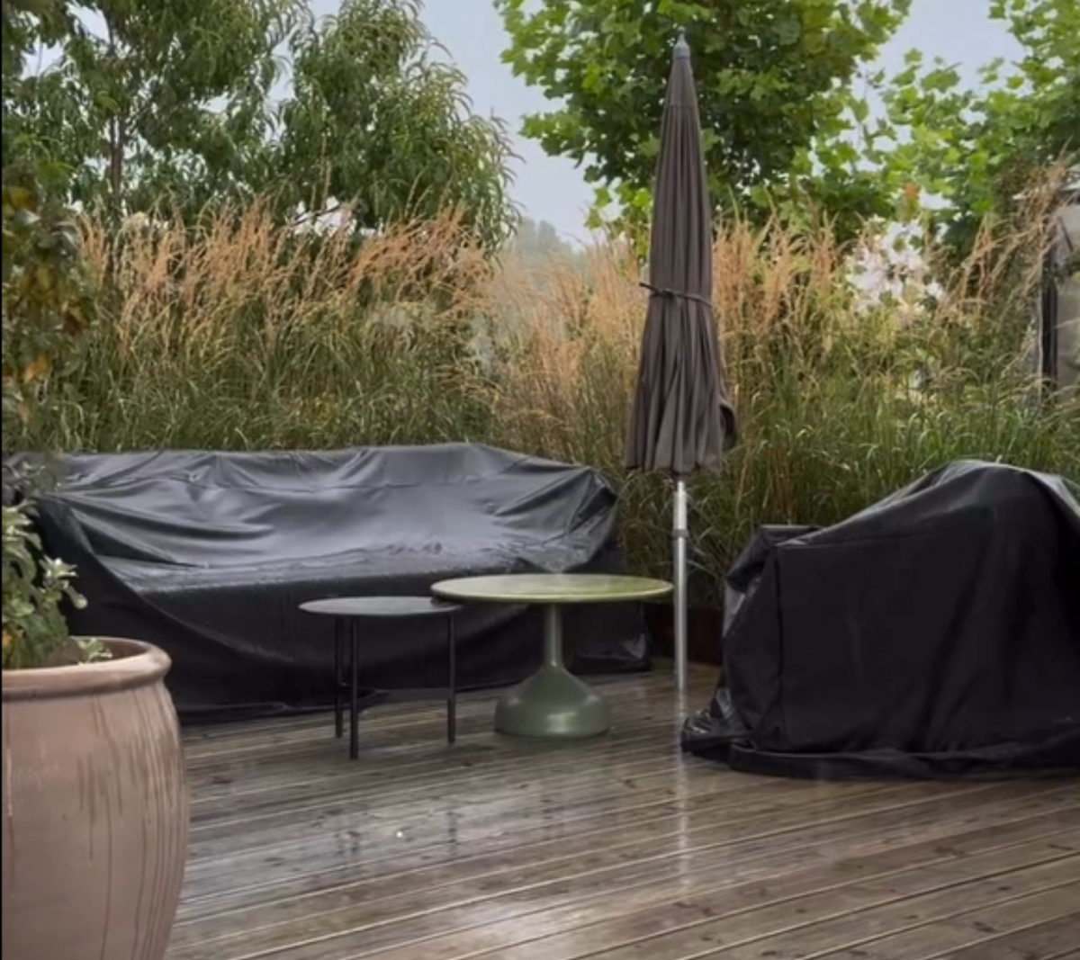 Außenbereich mit schwarzem Sofa, grünem Tisch und schwarzem Stuhl, umgeben von Pflanzen.