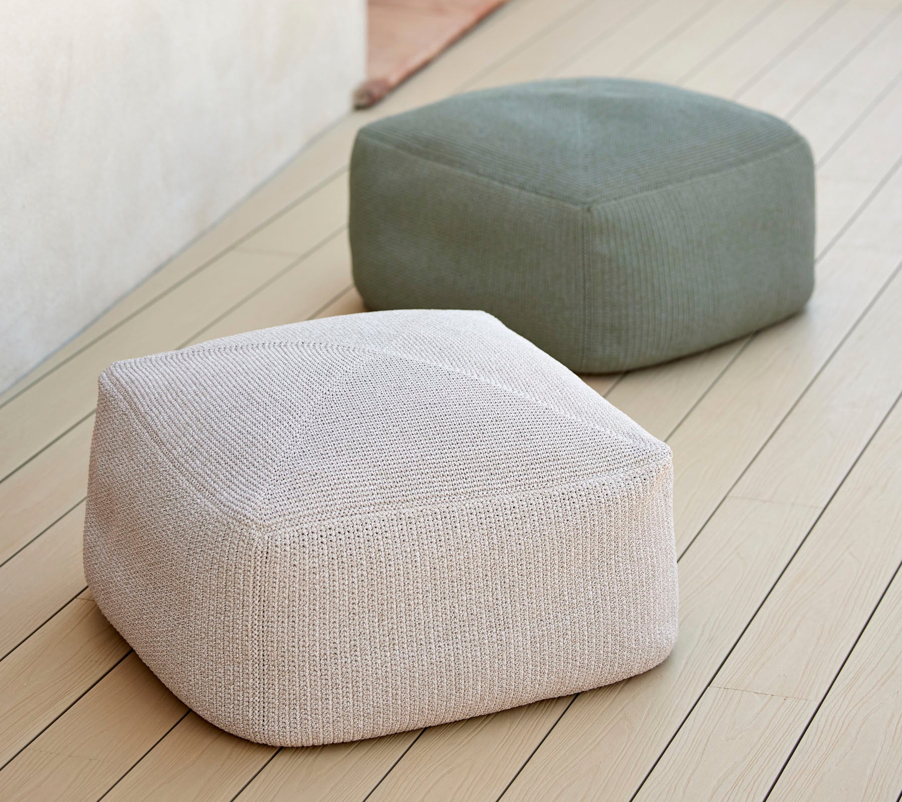 Zwei stilvolle, texturierte Poufs in neutralen Tönen, einer weiß und einer grün, auf einer glatten Oberfläche angeordnet, ideal für vielseitige Sitzgelegenheiten.