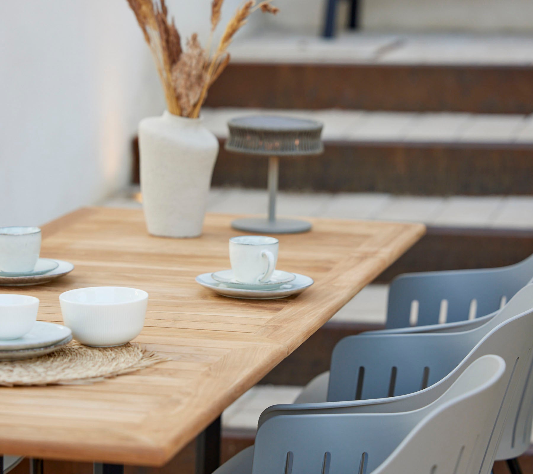 Ein Holztisch mit Tassen und Tellern gedeckt, dekoriert mit einer Vase mit getrockneten Blumen und grauen Stühlen in der Nähe.