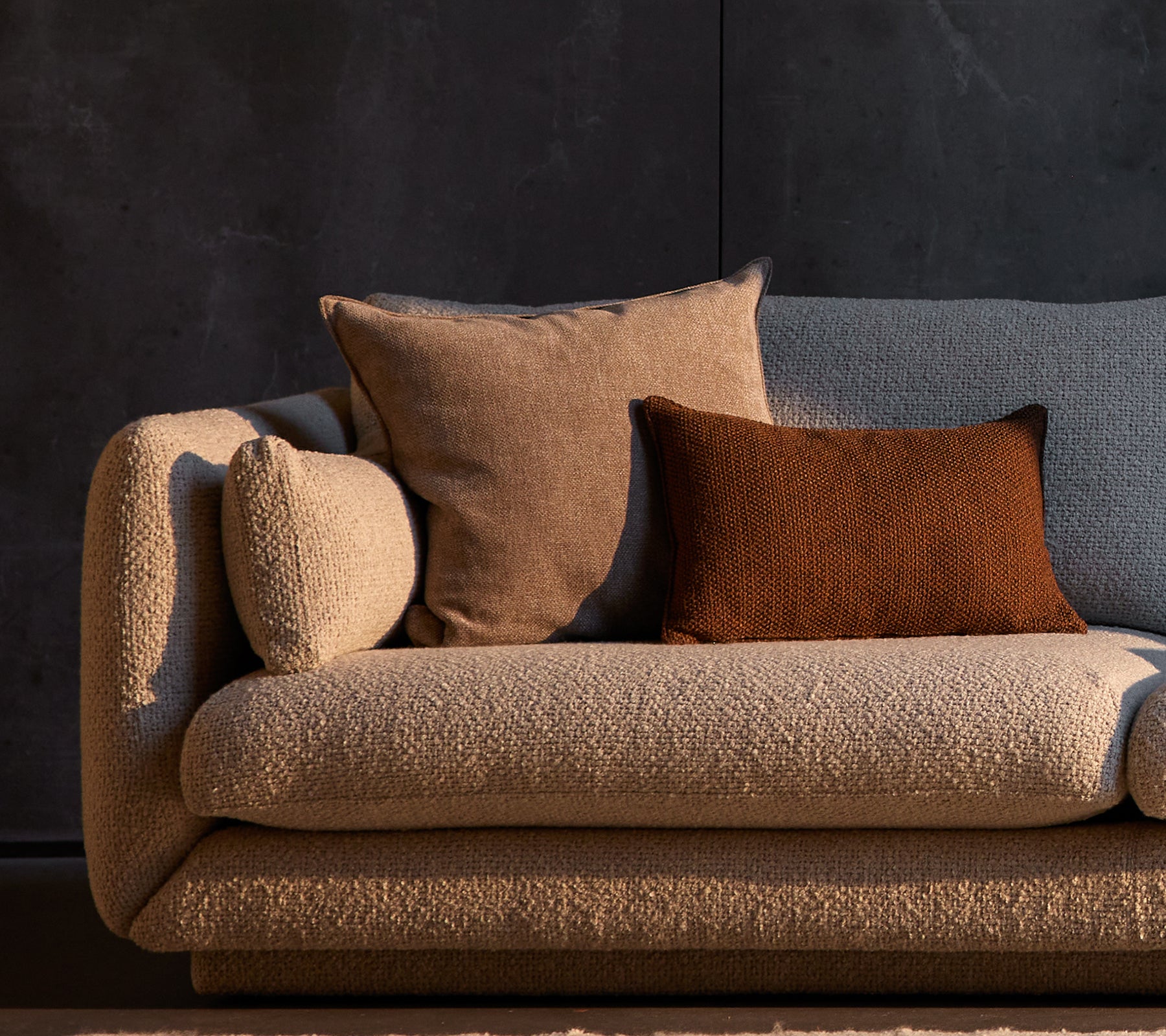 Essence- und Rise-Kissen auf einem strukturierten Bloom-Sofa, das neutrale Töne gegen einen dunklen Hintergrund zeigt.
