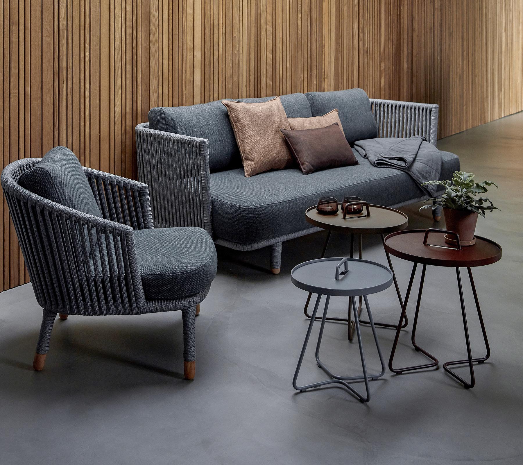 Stylishe Sitzanordnung mit einem grauen Sofa mit Kissen und zwei Akzentstühlen, begleitet von drei modernen Tischen und einer Topfpflanze.