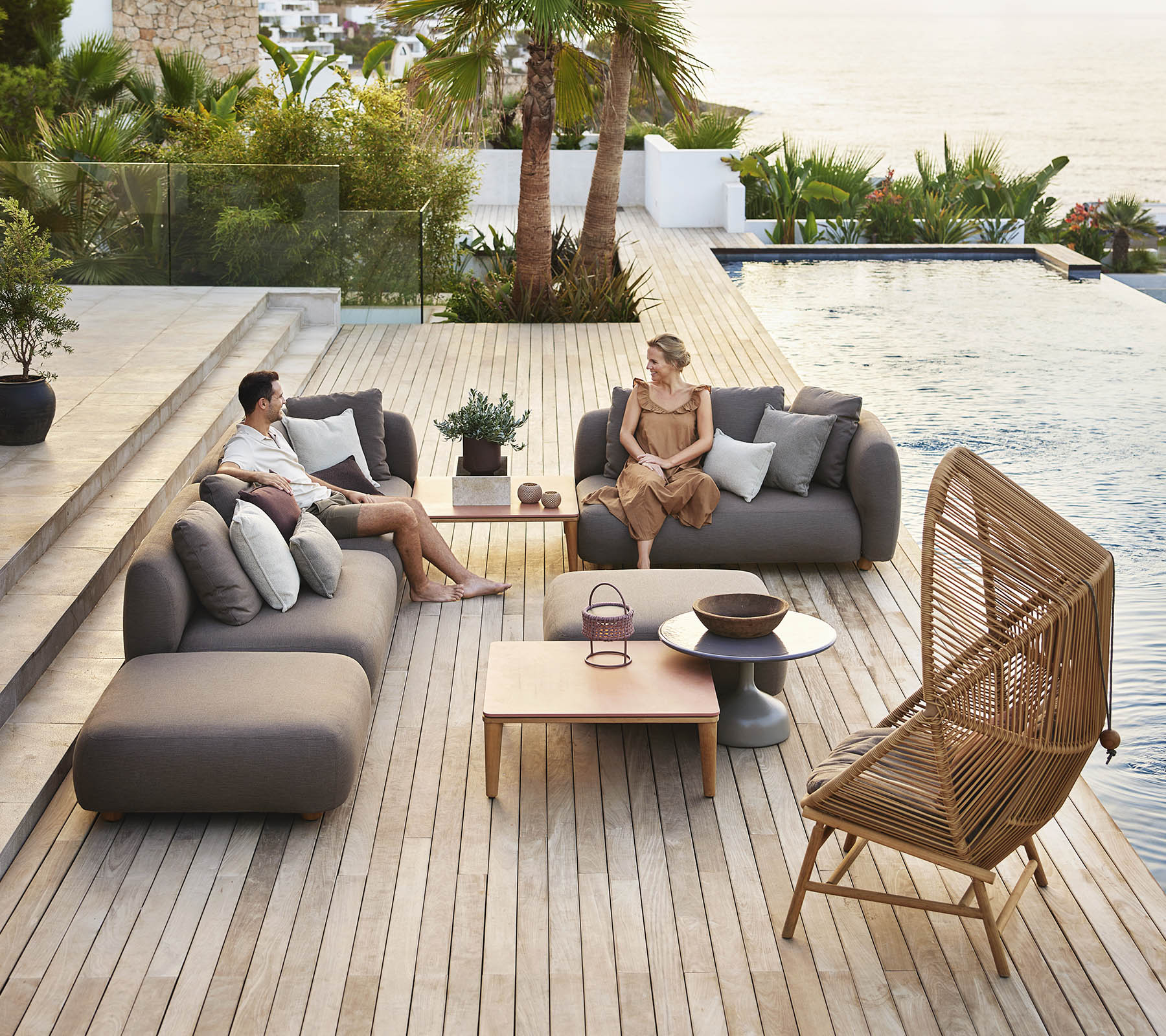 Zwei Personen entspannen sich auf modernen Sitzen am Pool, umgeben von Grün, mit einem Holztisch und dekorativen Pflanzen in der Nähe.