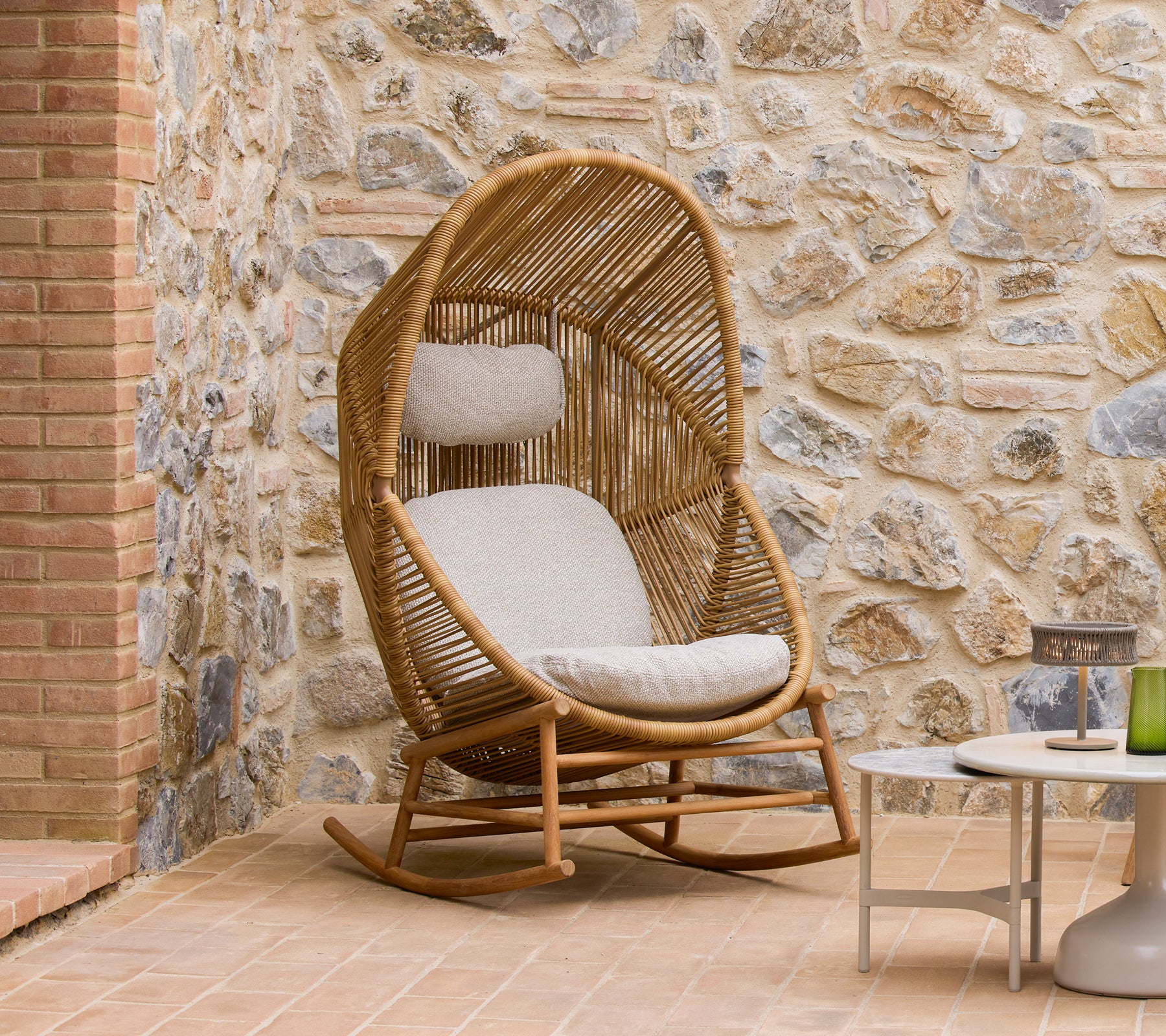 Hive Schaukelstuhl mit weichen Kissen neben Glaze-Couchtischen, vor einer strukturierten Steinwand.