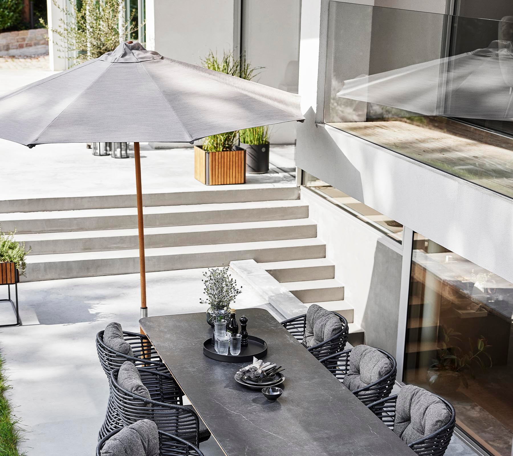 Ein stilvolles Essbereich im Freien mit einem langen Tisch, Korbstühlen und einem klassischen Sonnenschirm für Schatten.