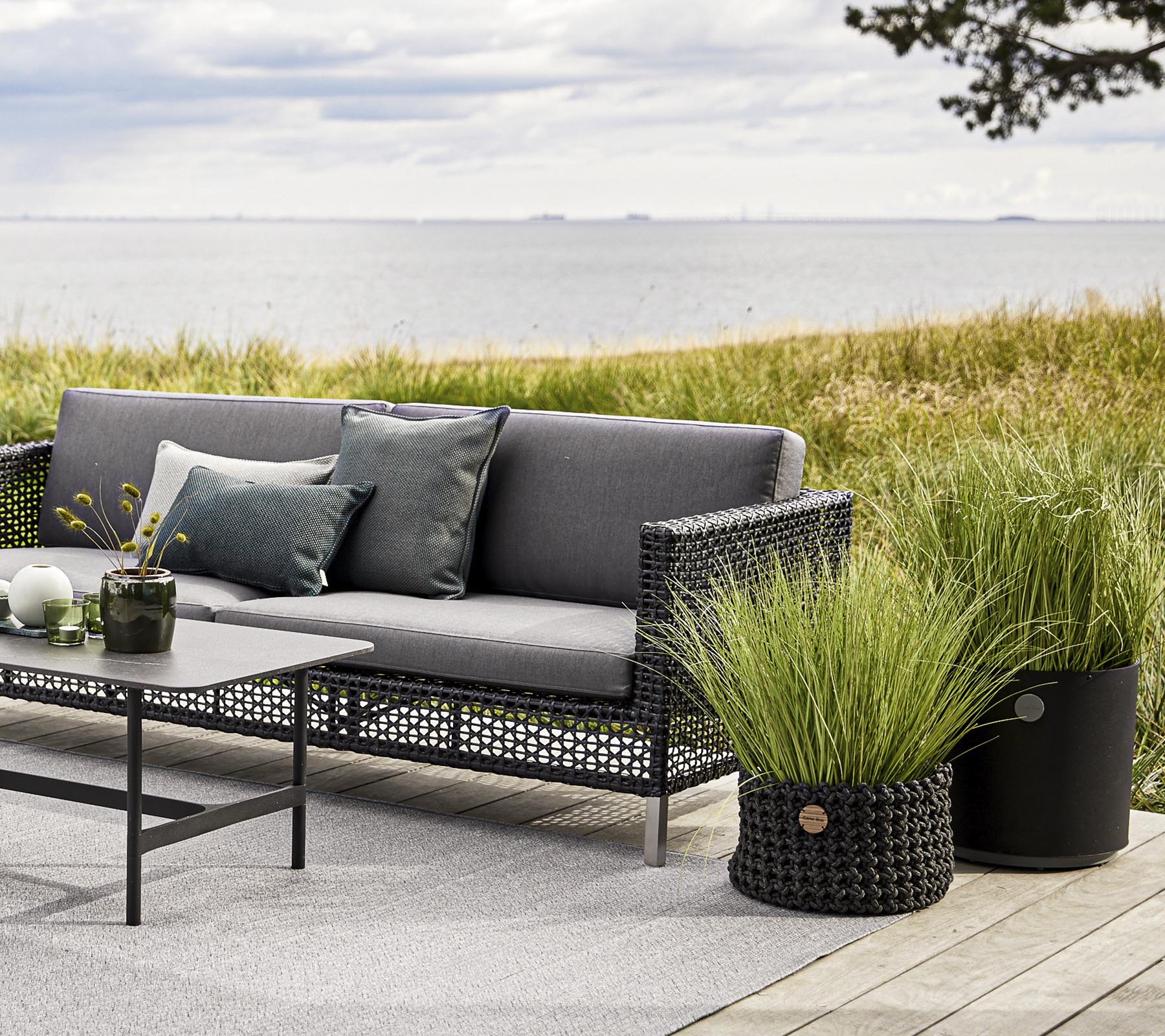 Stylische Außensitzgelegenheiten mit einem grauen Sofa, dekorativen Kissen und einem Tisch, ergänzt durch dekorative Grasblumentöpfe und eine ruhige Kulisse.