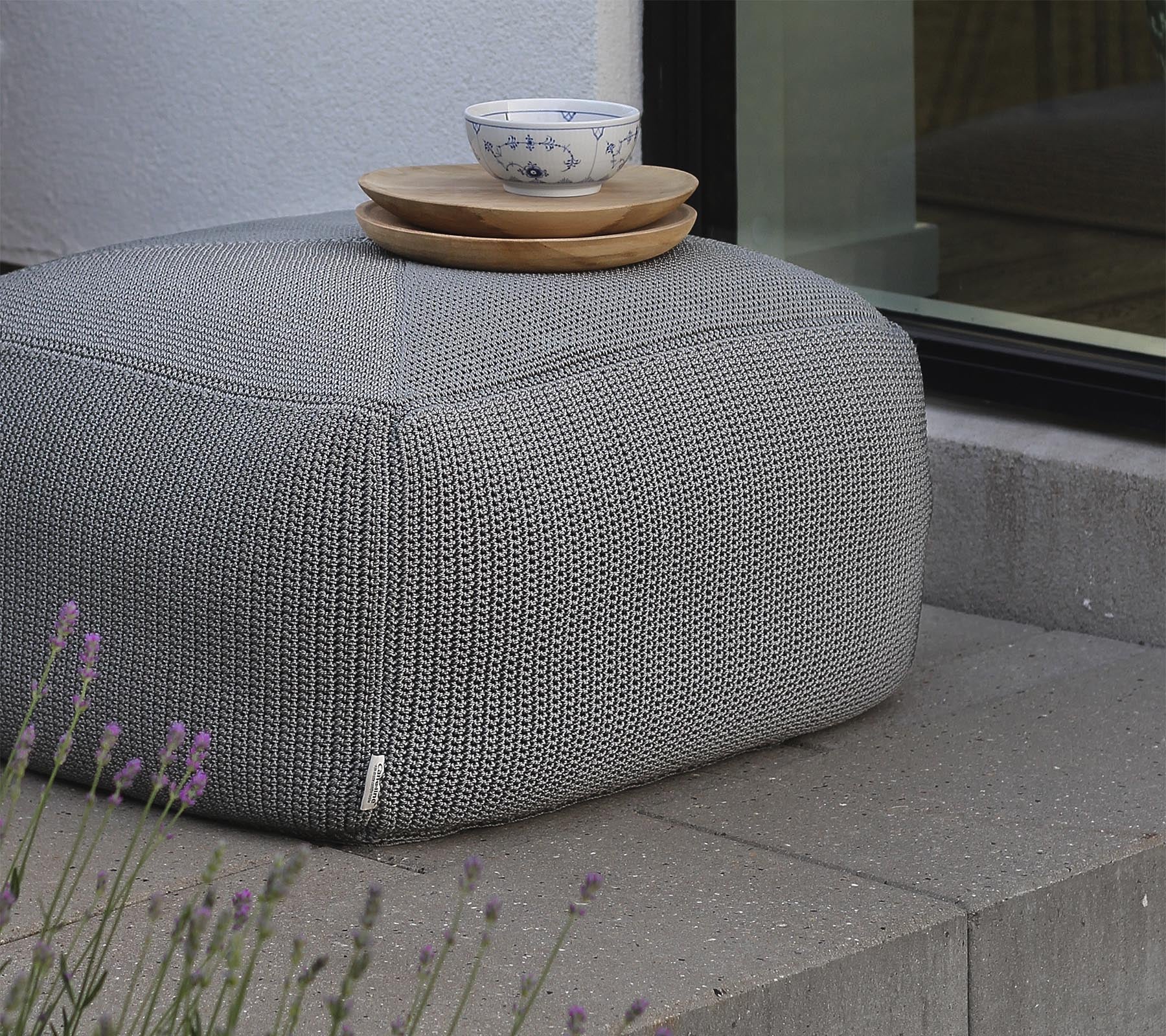 Ein strukturierter grauer Pouf, bedeckt mit Holzplatten und einer Keramikschüssel, positioniert in der Nähe eines Fensters und umgeben von Lavendel.