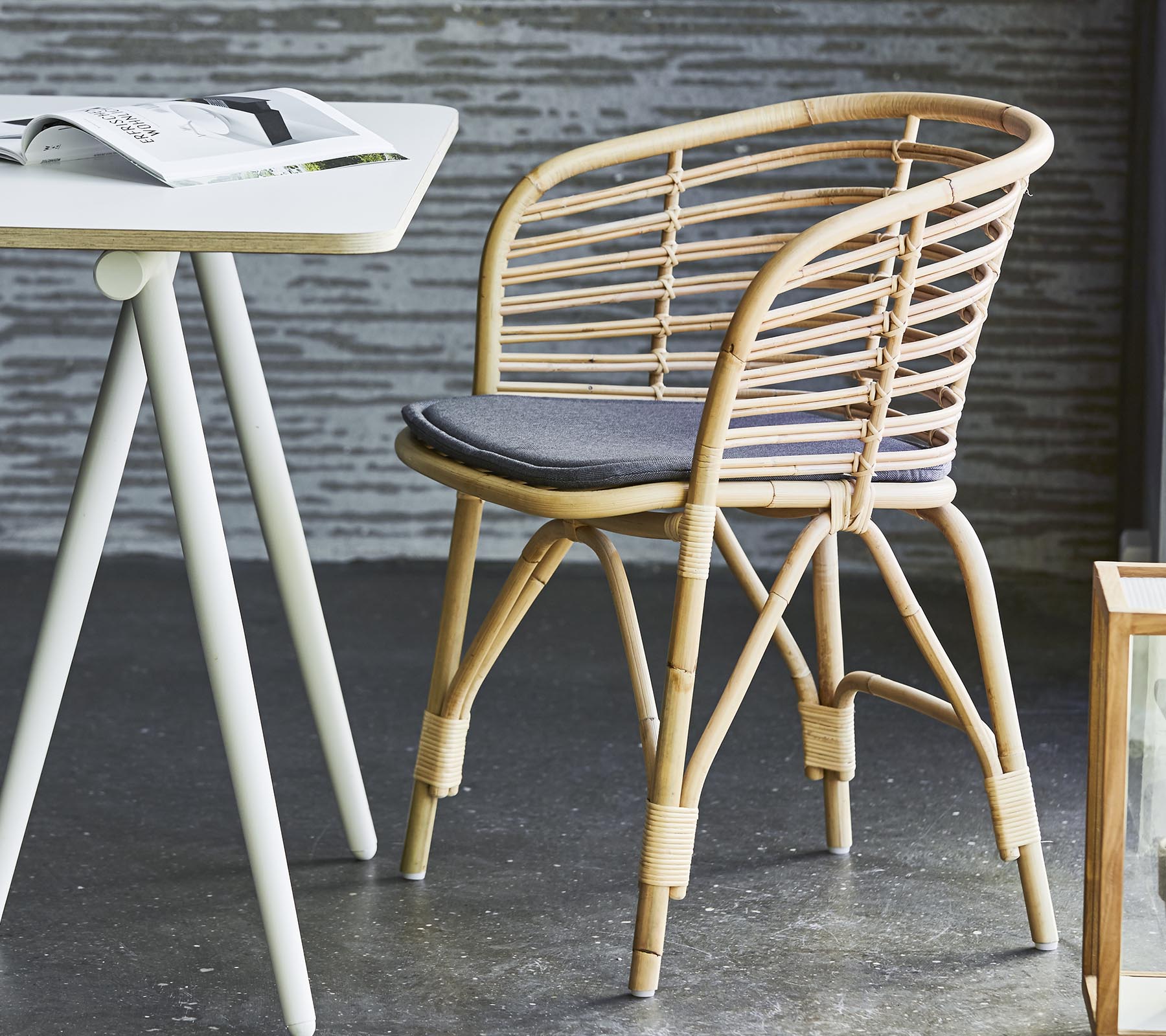 Stylischer Rattanstuhl mit einem grauen Kissen, kombiniert mit einem schlichten Tisch und zeitgenössischer Dekoration. Geeignet für legere Sitzgelegenheiten und Entspannung.