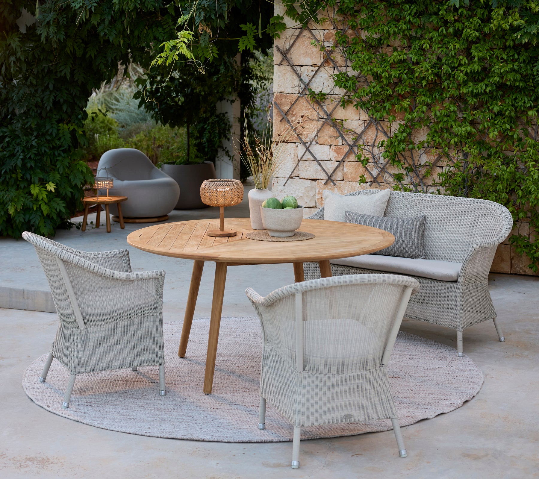 Moderne Lounge-Bereich mit grauen Stühlen, einem runden Holztisch und einem grauen Sofa, umgeben von Grün.