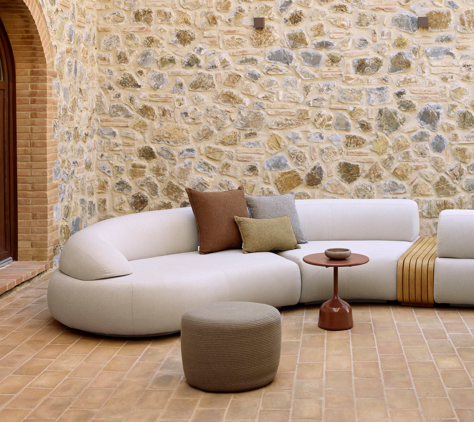 Gemütlicher Loungebereich mit einem geschwungenen Sofa, einem Glaze-Couchtisch und einem runden Fußhocker vor einer strukturierten Steinwand.