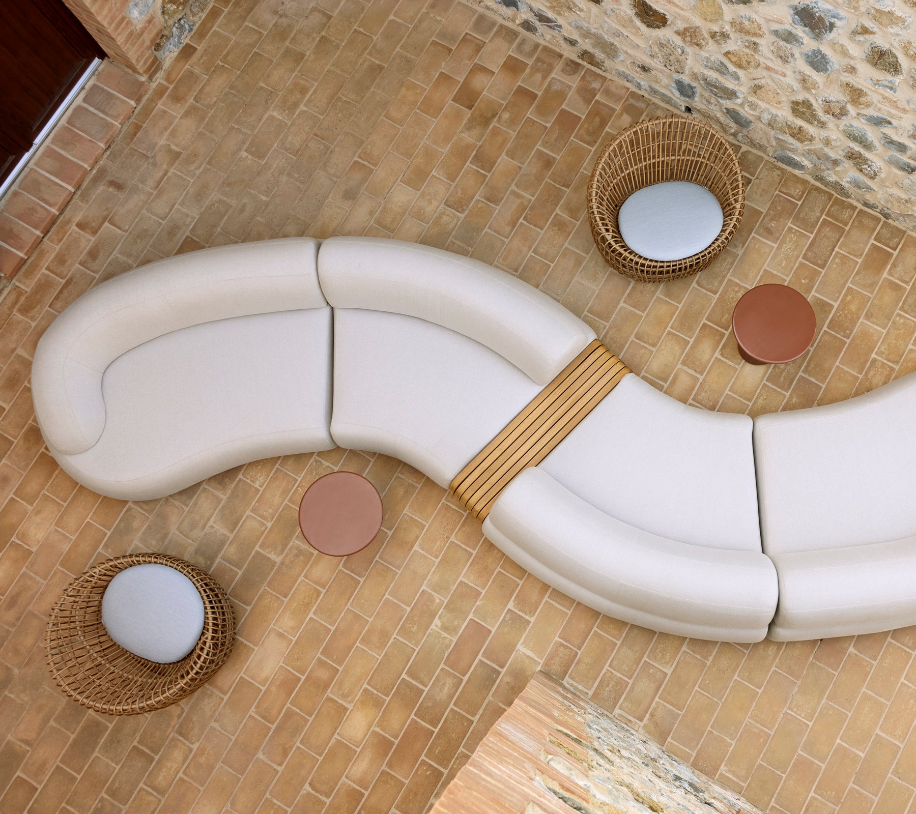 Gemütliche Lounge mit einer geschwungenen weißen Sitzgelegenheit, zwei runden Glaze- couchtischen und geflochtenen Nest-Stühlen auf einem Steinfussboden.