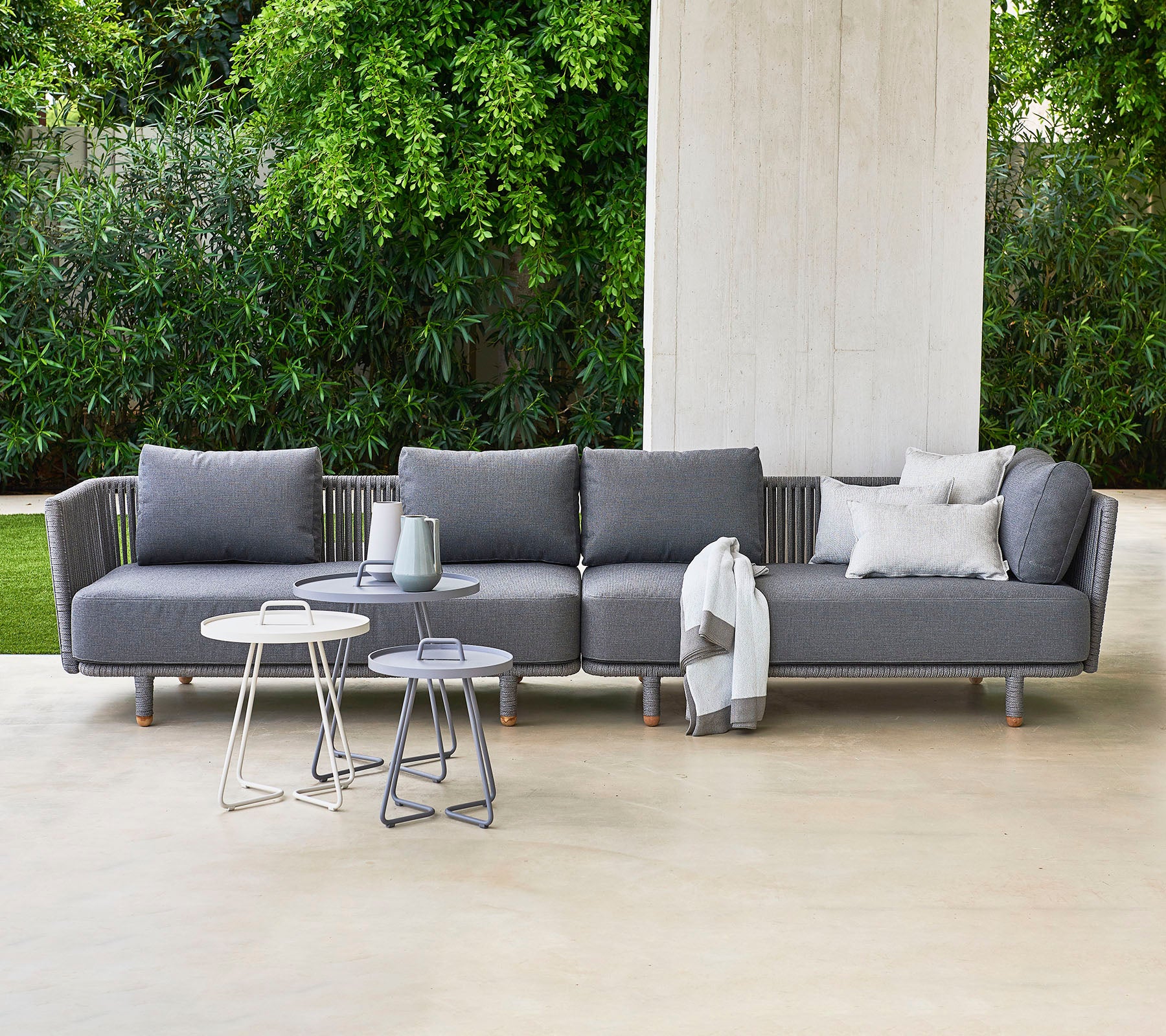 Moderne graues Ecksofa mit Kissen, begleitet von zwei stilvollen Beistelltischen, vor einem Hintergrund aus üppigem Grün.