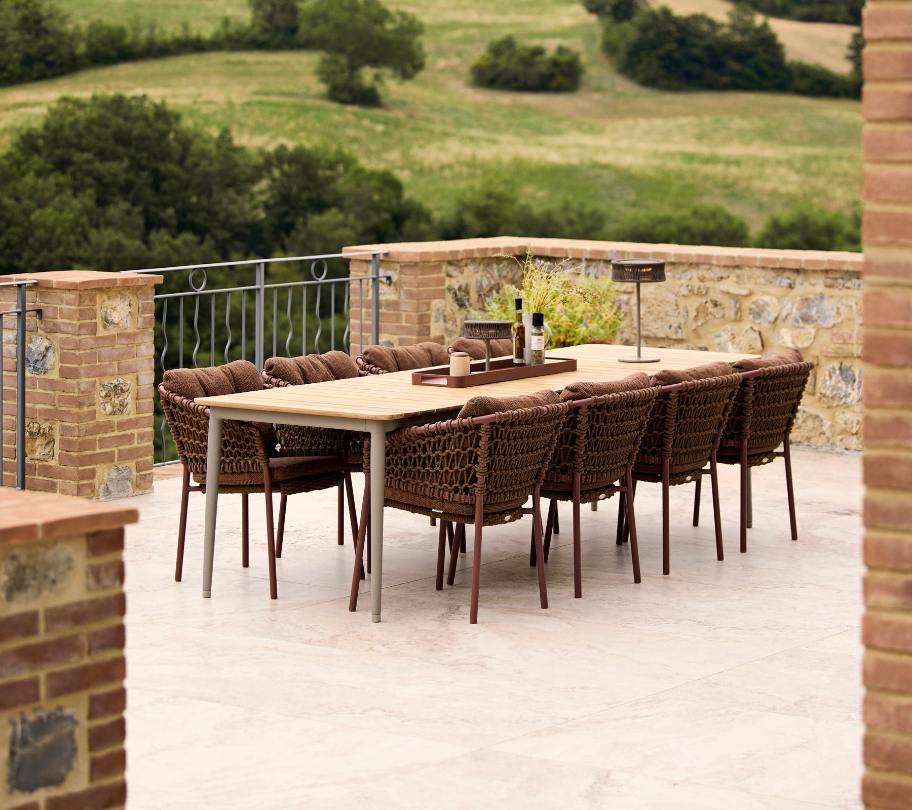 Umberbraune Ozeanstühle umgeben einen Core Esstisch, vor einer üppig grünen Landschaft. Elegante Outdoor-Dining-Anordnung.