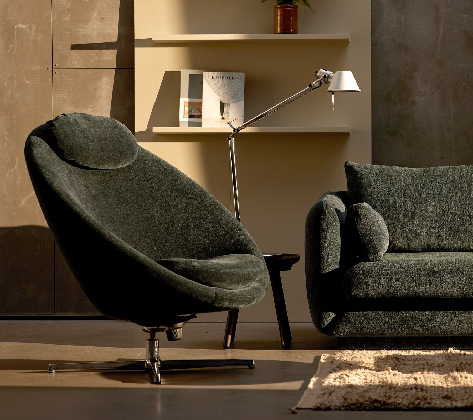Pace-Stuhl aus dunkelgrüner Chenille neben einem Bloom-Sofa, ergänzt durch eine elegante Lampe und minimalistische Regaldekoration.