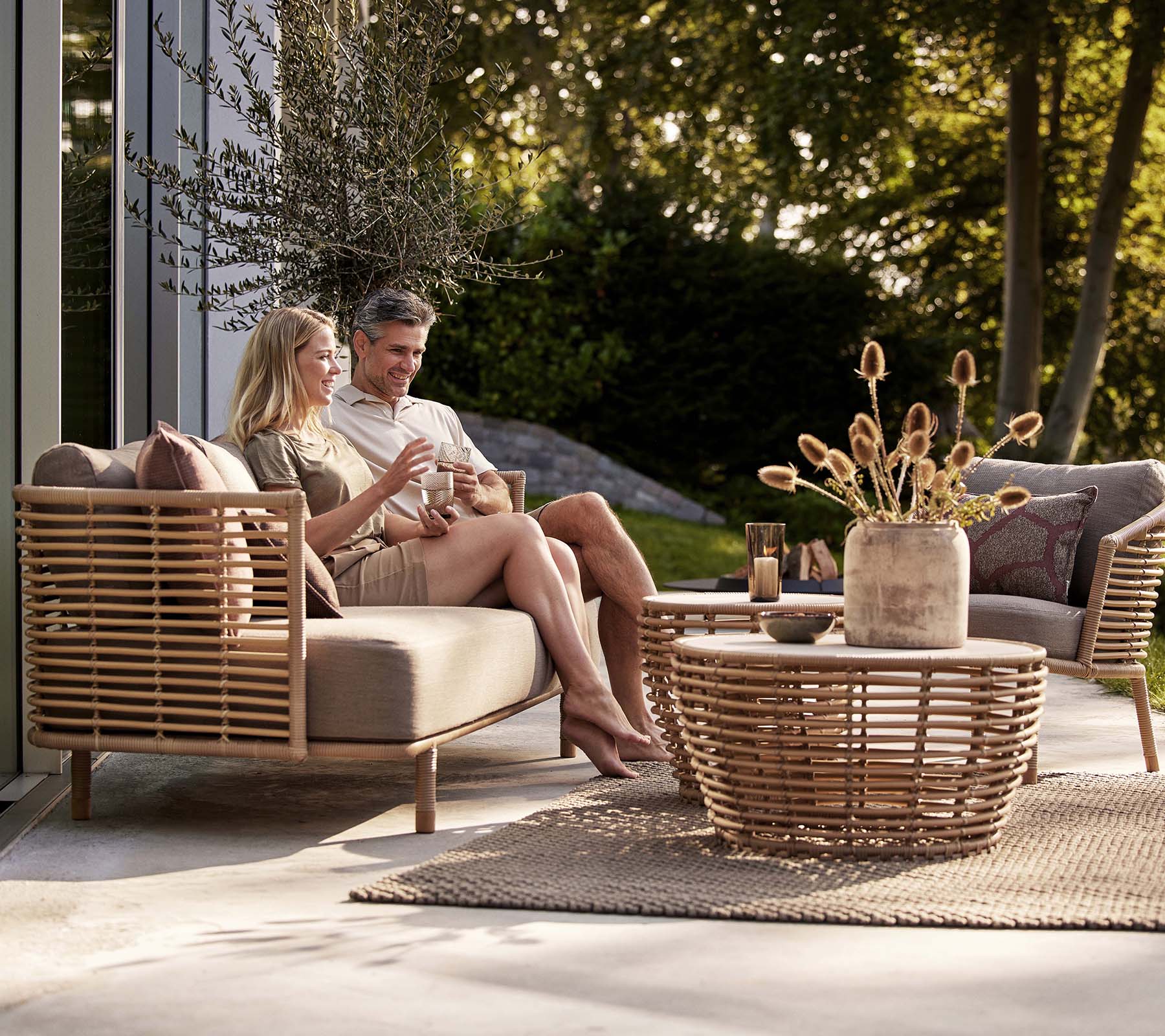 Ein Paar genießt Getränke auf einem Sense-Sofa und einem Loungesessel, begleitet von einem stilvollen Korbkaffee-Tisch und einer dekorativen Vase.