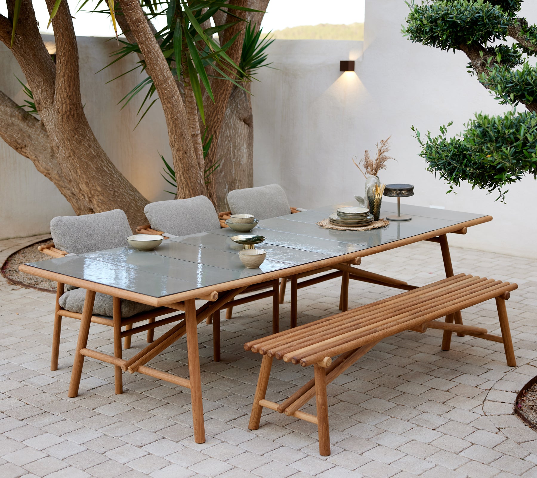 Teak-Esstisch mit Glasplatte, begleitet von einer Bank und vier gepolsterten Stühlen, gedeckt mit Geschirr und atmosphärischer Beleuchtung.