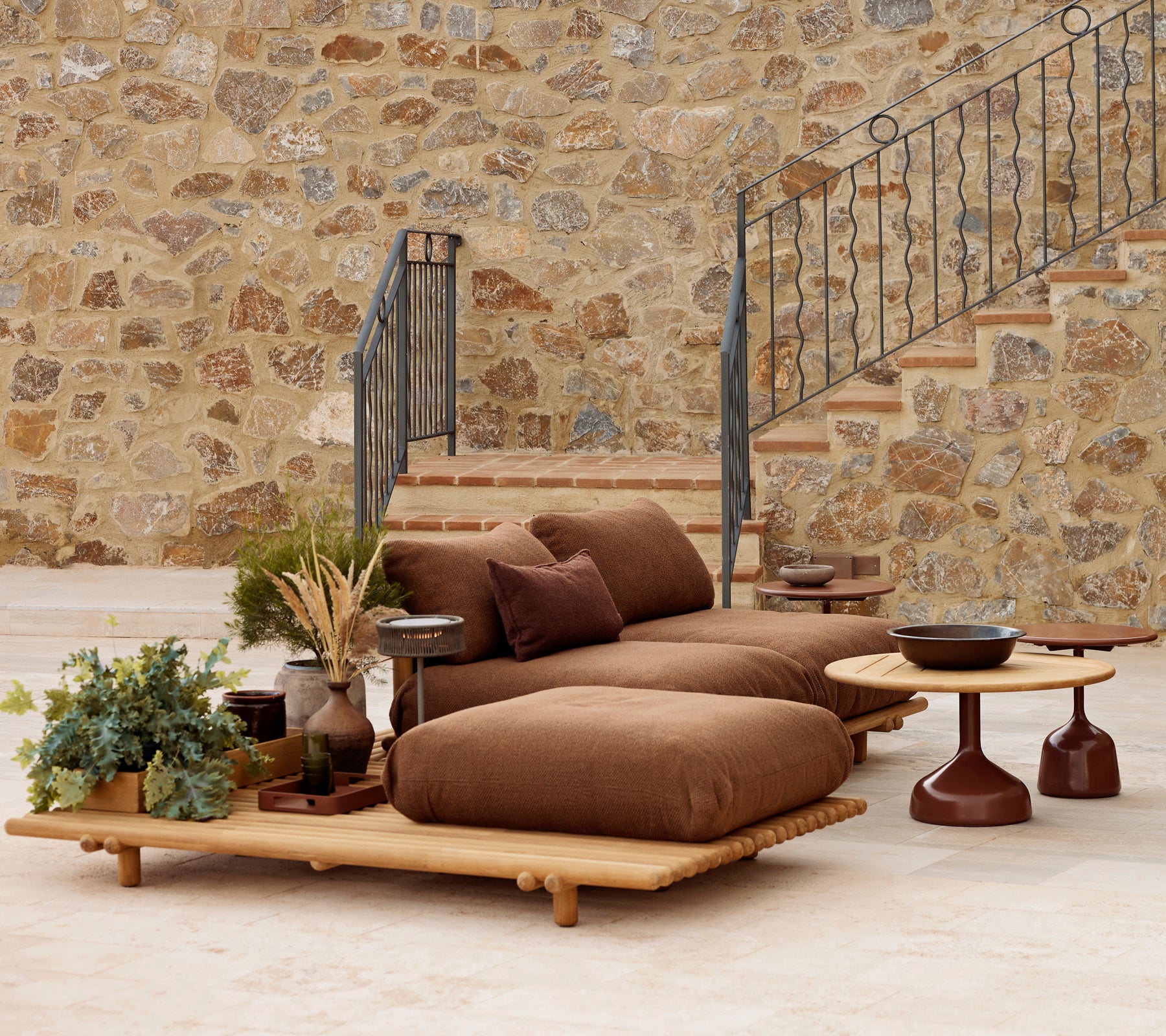 Teak-Lounge mit plüschigen Kissen und einem Glaze-Couchtisch, ergänzt durch dekorative Pflanzen und eine strukturierte Steinwand.