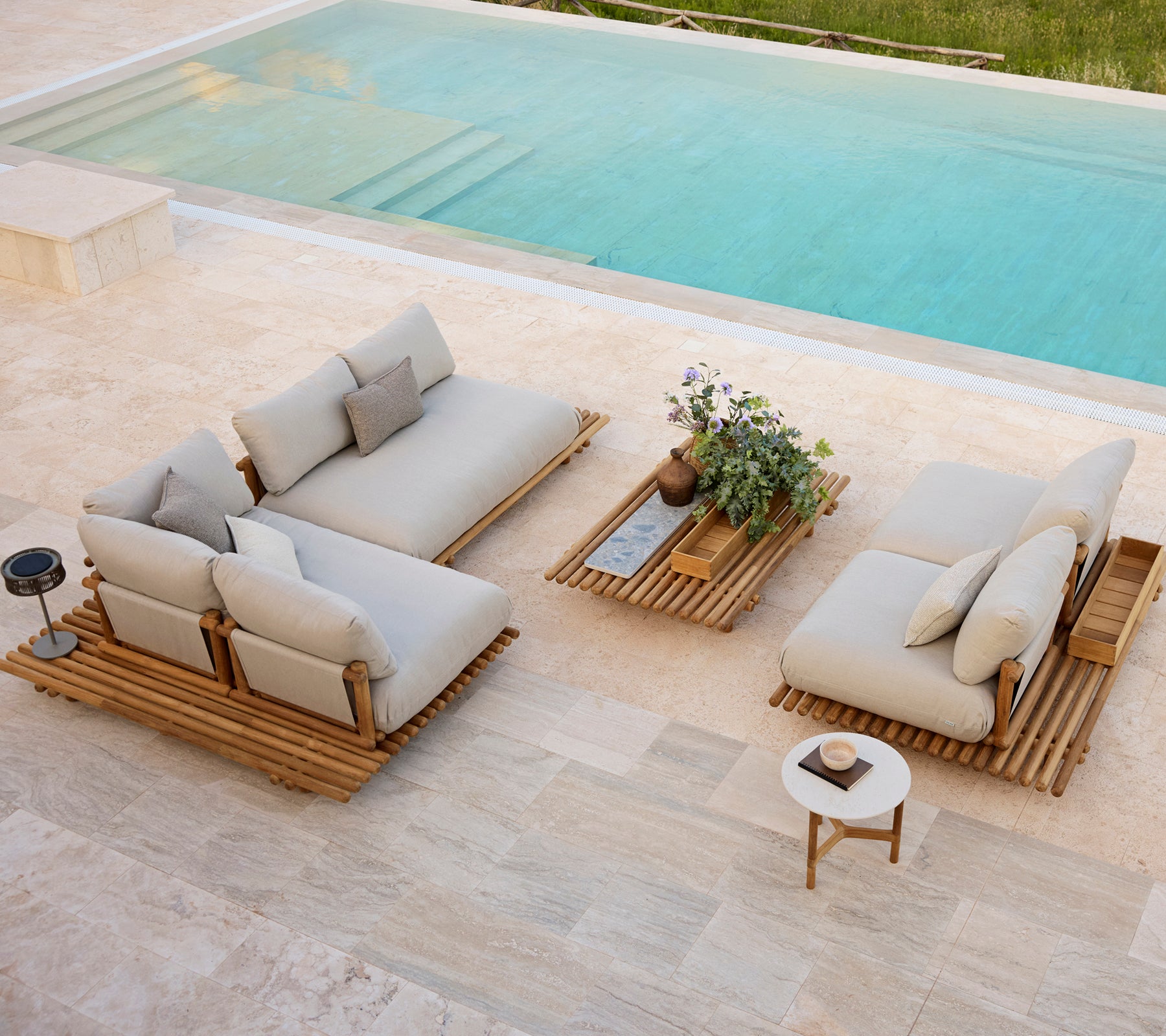 Moderne Außenbereich mit neutralen Kissen, Holzplattform-Tischen und einer kleinen Topfpflanze neben einem Pool.