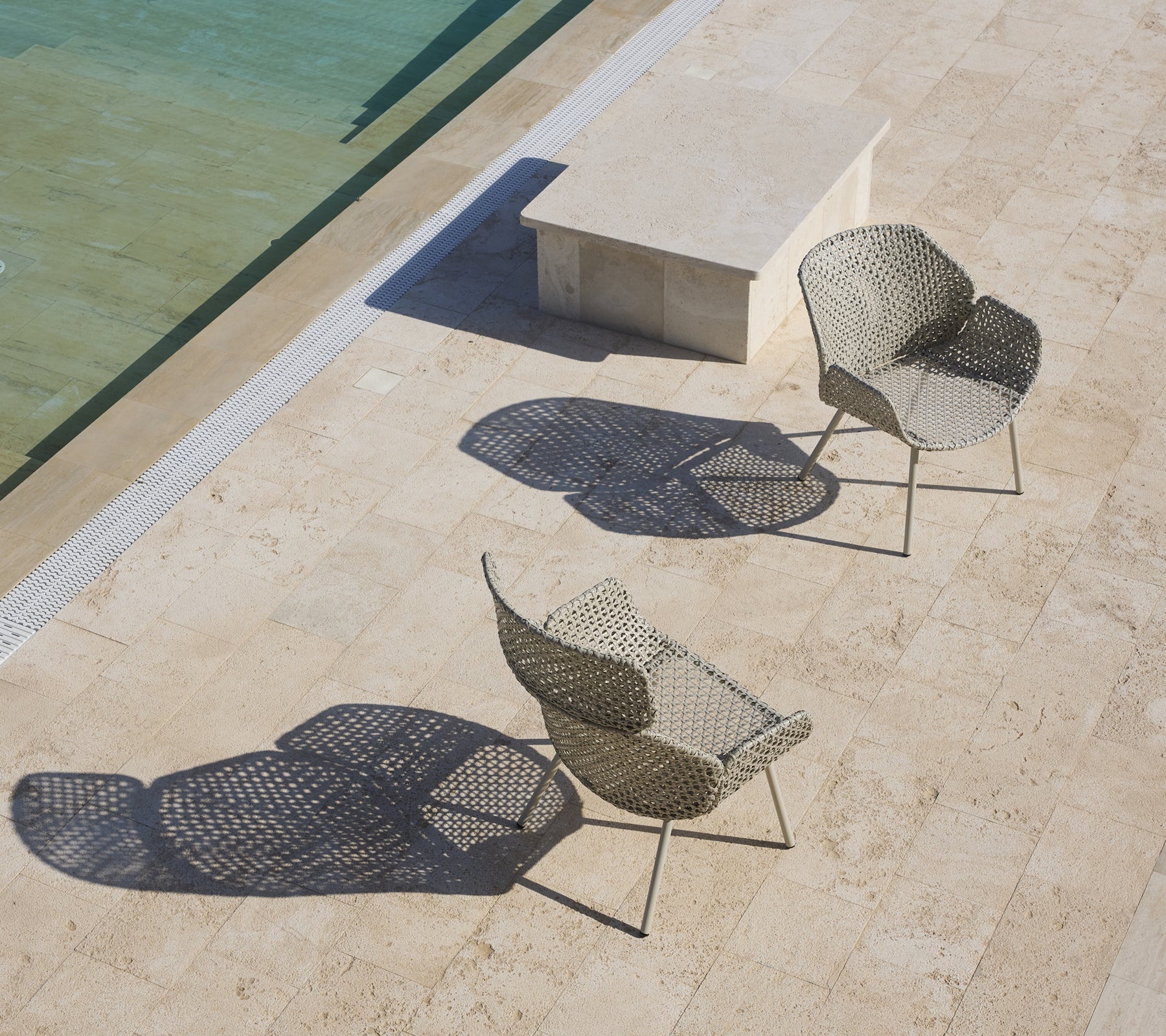 Zwei stilvolle Lounge-Stühle mit einzigartigen Designs, die Schatten werfen, positioniert in der Nähe einer ruhigen Wasseroberfläche und einem Steintisch.