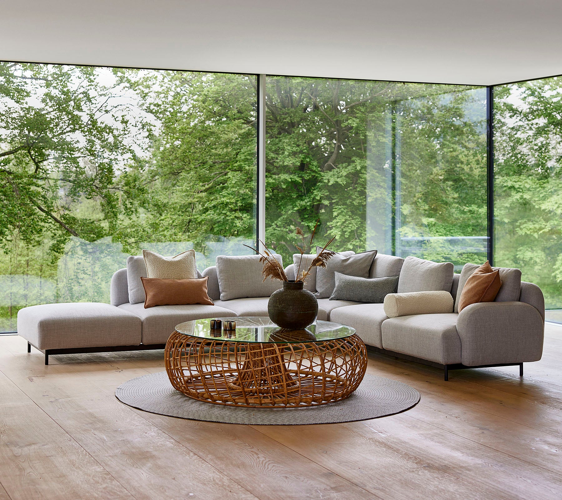 Moderner Lounge-Bereich mit einem modularen Sofa mit dekorativen Kissen, einem runden, geflochtenen Couchtisch und großen Fenstern, die Grünflächen zeigen.