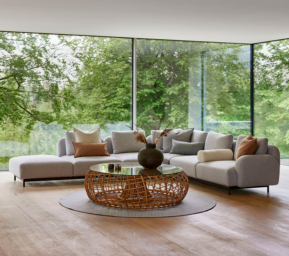Moderner Lounge-Bereich mit einem modularen Sofa mit dekorativen Kissen, einem runden, geflochtenen Couchtisch und großen Fenstern, die Grünflächen zeigen.