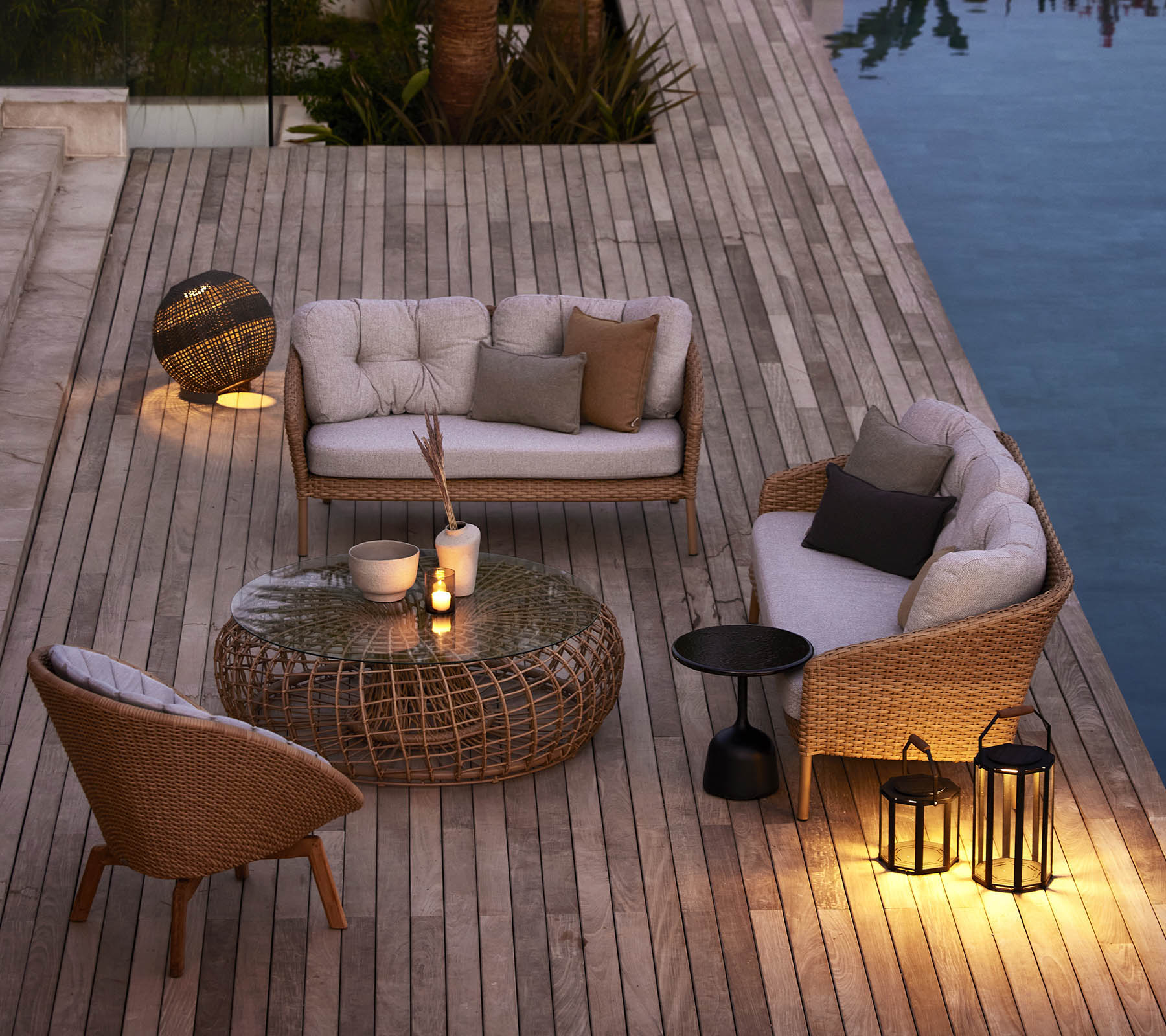 Gemütlicher Außenbereich mit geflochtenen Möbeln, weichen Kissen und ambienter Beleuchtung neben einem Pool auf einer Holzterrasse.