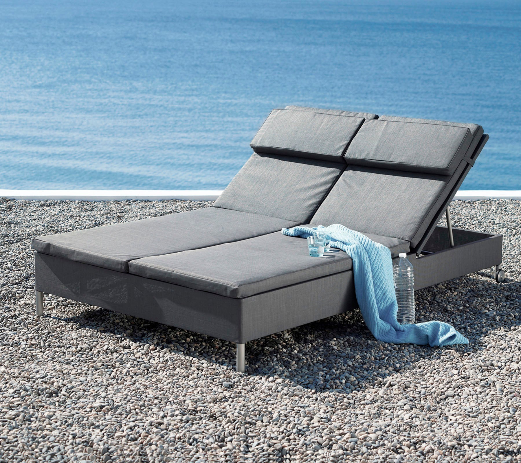 Ein stilvoller grauer Loungesessel mit verstellbarem Rückenlehne, accompanied by a weichen Überwurf, neben einer ruhigen Wasseransicht.